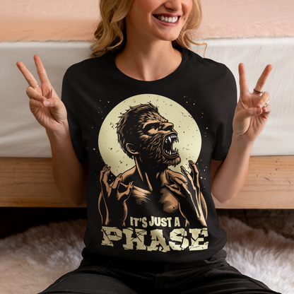 This t-shirt features a dynamic illustration of a man mid-transformation into a fearsome werewolf under the glow of a full moon. His human features contort as claws emerge, fur spreads across his body, and his eyes glow with a supernatural intensity. Below the image, the humorous and clever text reads: "IT'S JUST A PHASE," playfully alluding to both the lunar cycle and the werewolf's transformation.