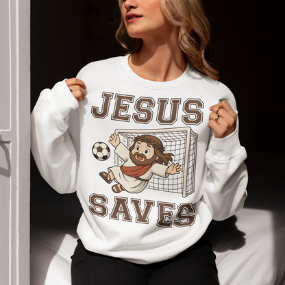 White sweatshirt featuring a cartoon of Jesus as a soccer goalie diving to block a ball in front of a goal net, with the bold text “Jesus Saves” above and below. Fun, humorous Christian design combining faith and sports, printed on a clean white background.