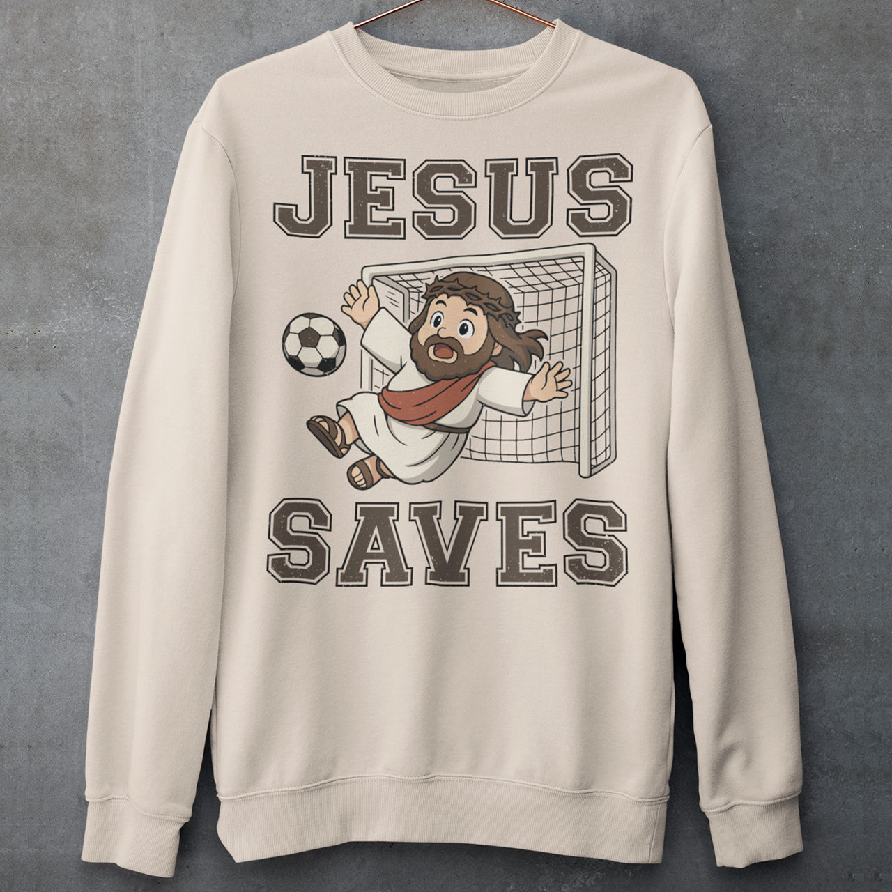 Beige sweatshirt featuring a cartoon of Jesus as a soccer goalie diving to block a ball in front of a goal net, with the bold text “Jesus Saves” above and below. Fun, humorous Christian design combining faith and sports, printed on a clean white background.