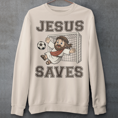 Beige sweatshirt featuring a cartoon of Jesus as a soccer goalie diving to block a ball in front of a goal net, with the bold text “Jesus Saves” above and below. Fun, humorous Christian design combining faith and sports, printed on a clean white background.