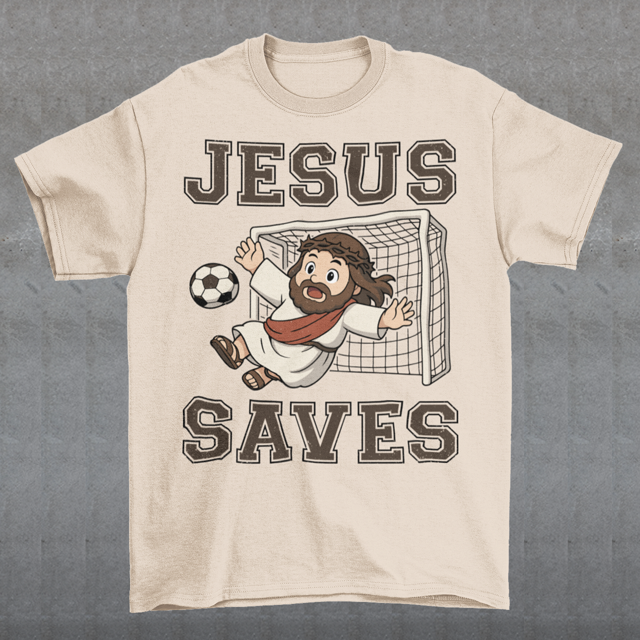 Beige t-shirt featuring a cartoon of Jesus as a soccer goalie diving to block a ball in front of a goal net, with the bold text “Jesus Saves” above and below. Fun, humorous Christian design combining faith and sports, printed on a clean white background.