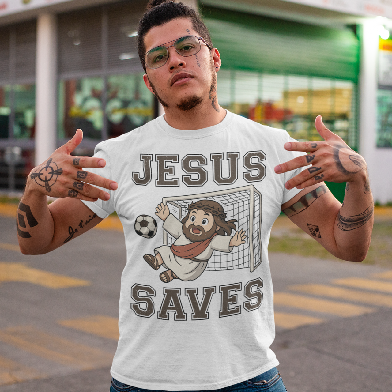 White t-shirt featuring a cartoon of Jesus as a soccer goalie diving to block a ball in front of a goal net, with the bold text “Jesus Saves” above and below. Fun, humorous Christian design combining faith and sports, printed on a clean white background.