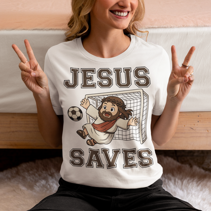 White t-shirt featuring a cartoon of Jesus as a soccer goalie diving to block a ball in front of a goal net, with the bold text “Jesus Saves” above and below. Fun, humorous Christian design combining faith and sports, printed on a clean white background.