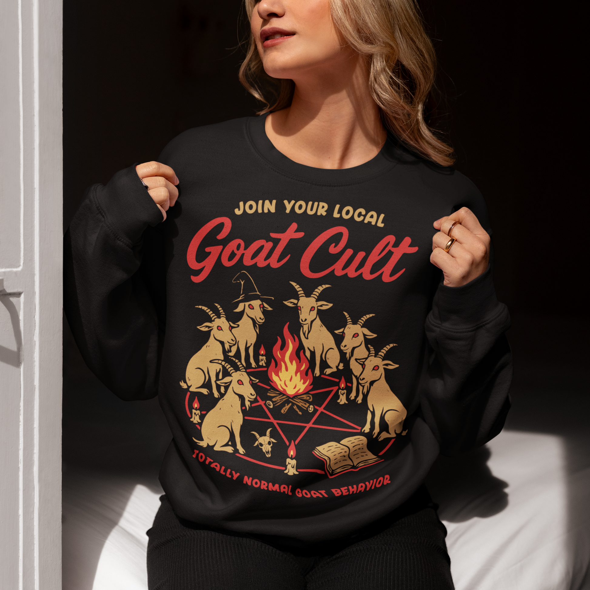 Black sweatshirt featuring a humorous occult-style graphic of goats gathered in a ritual circle around a fire with candles and symbols. Text reads “Join Your Local Goat Cult” with “Totally Normal Goat Behavior” underneath. The design has a playful spooky aesthetic with cartoon goats, making it a funny and quirky dark humor piece.