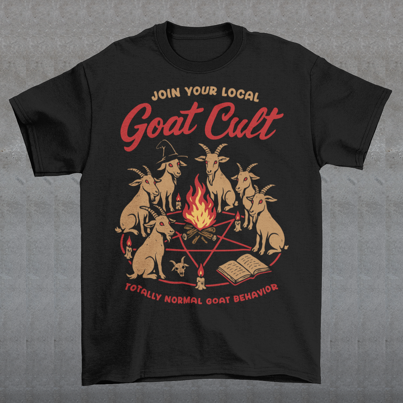 Black t-shirt featuring a humorous occult-style graphic of goats gathered in a ritual circle around a fire with candles and symbols. Text reads “Join Your Local Goat Cult” with “Totally Normal Goat Behavior” underneath. The design has a playful spooky aesthetic with cartoon goats, making it a funny and quirky dark humor piece.