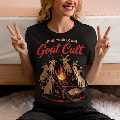 Black t-shirt featuring a humorous occult-style graphic of goats gathered in a ritual circle around a fire with candles and symbols. Text reads “Join Your Local Goat Cult” with “Totally Normal Goat Behavior” underneath. The design has a playful spooky aesthetic with cartoon goats, making it a funny and quirky dark humor piece.