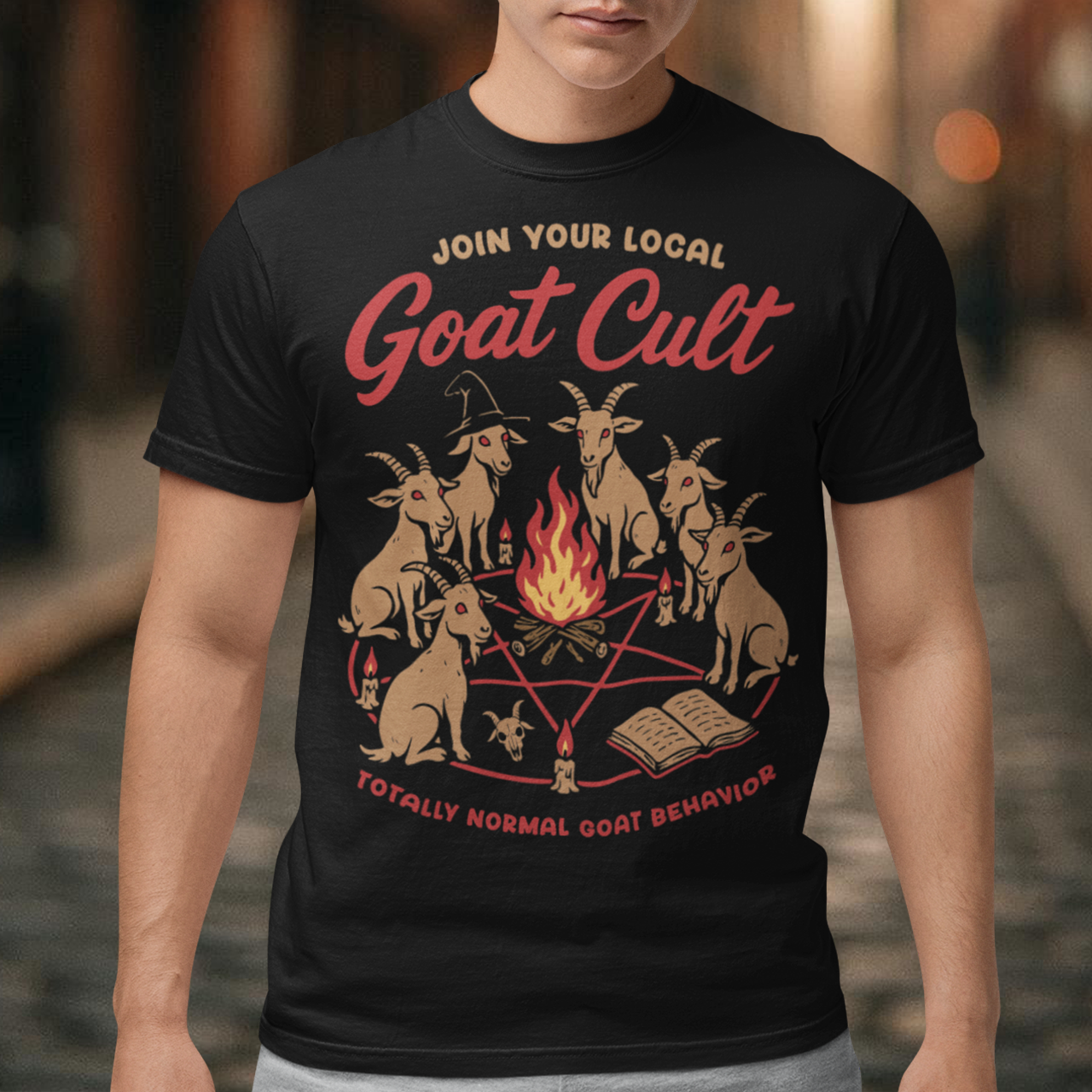 Black t-shirt featuring a humorous occult-style graphic of goats gathered in a ritual circle around a fire with candles and symbols. Text reads “Join Your Local Goat Cult” with “Totally Normal Goat Behavior” underneath. The design has a playful spooky aesthetic with cartoon goats, making it a funny and quirky dark humor piece.