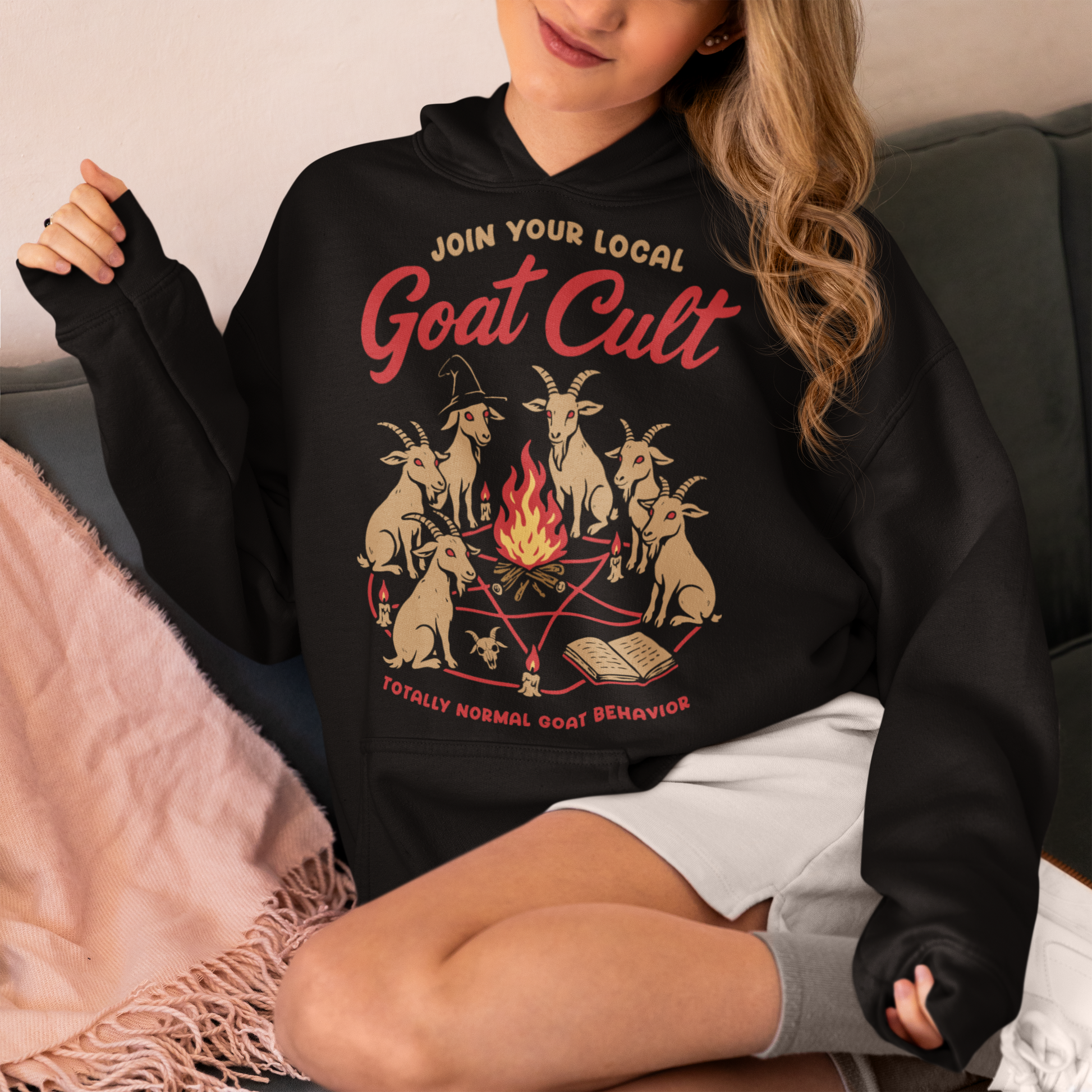 Black hoodie featuring a humorous occult-style graphic of goats gathered in a ritual circle around a fire with candles and symbols. Text reads “Join Your Local Goat Cult” with “Totally Normal Goat Behavior” underneath. The design has a playful spooky aesthetic with cartoon goats, making it a funny and quirky dark humor piece.