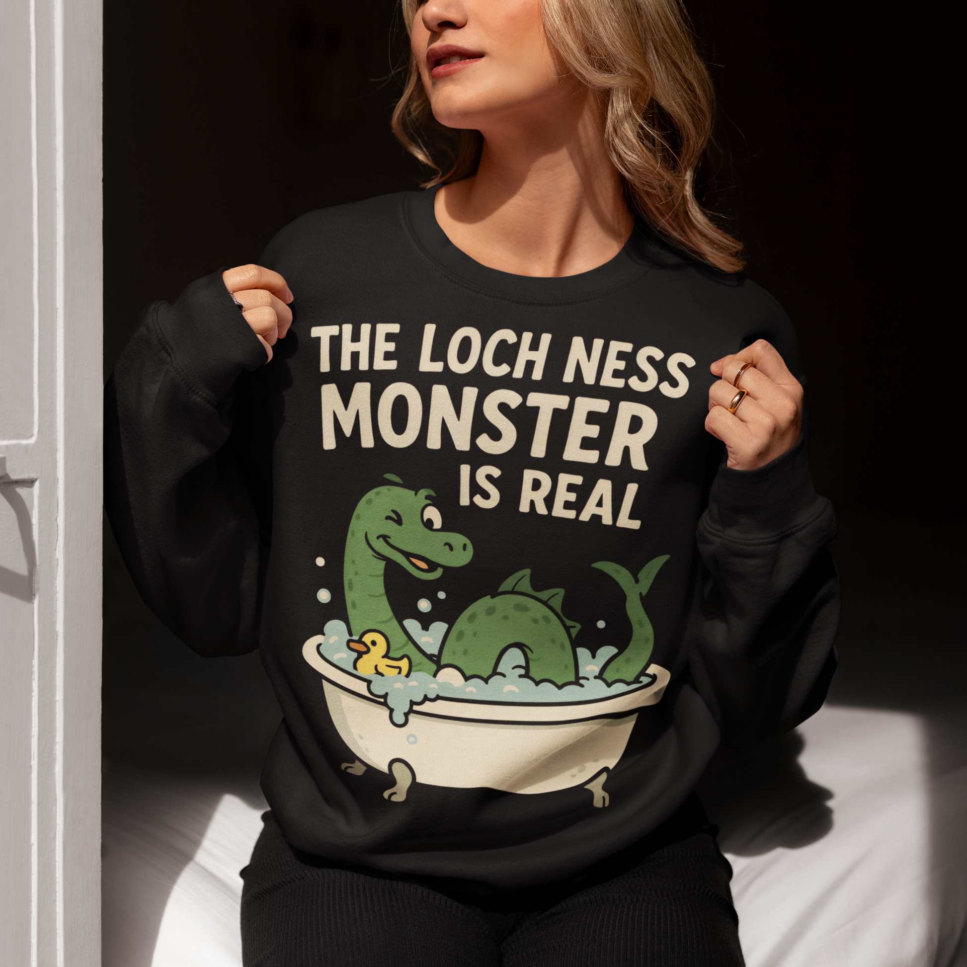 This black graphic sweatshirt puts a playful spin on legendary folklore with an adorable take on the Loch Ness Monster. Featuring a cheerful green Nessie splashing in a clawfoot bathtub with bubbles and a rubber duck, the design pairs cozy humor with cryptid charm. Bold lettering declares “The Loch Ness Monster Is Real,” delivering the punchline with confidence and a wink.