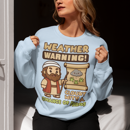 Light Blue sweatshirt featuring a cute cartoon Moses pointing at a weather scroll that shows a smiling rain cloud and falling frogs. The text reads “Weather Warning! Cloudy With a Chance of Frogs.” The design uses playful retro colors with funny biblical humor inspired by the plague of frogs story.