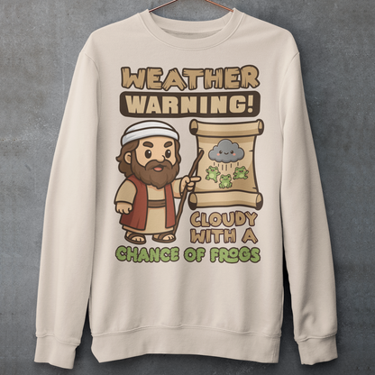 Beige sweatshirt featuring a cute cartoon Moses pointing at a weather scroll that shows a smiling rain cloud and falling frogs. The text reads “Weather Warning! Cloudy With a Chance of Frogs.” The design uses playful retro colors with funny biblical humor inspired by the plague of frogs story.