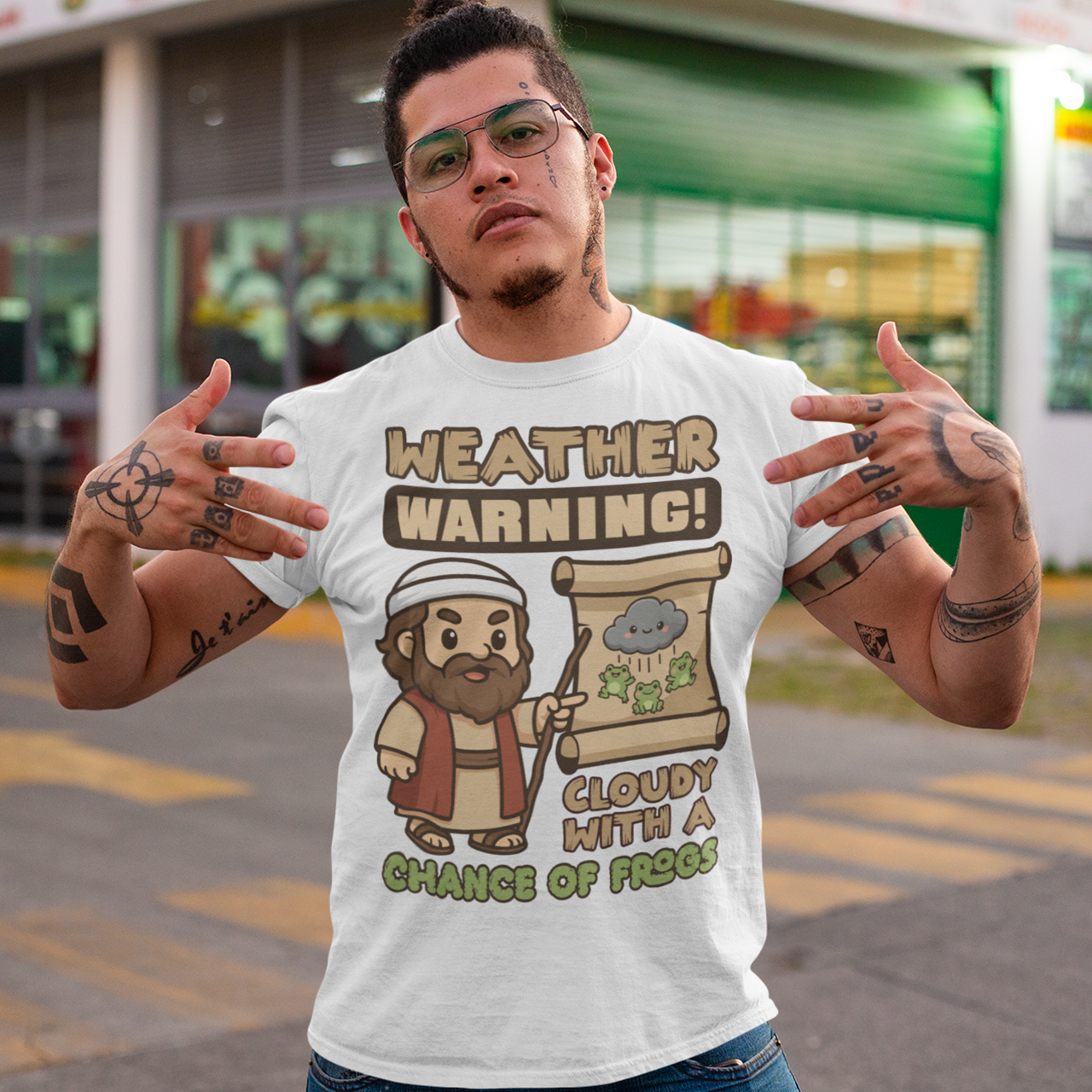 White t-shirt featuring a cute cartoon Moses pointing at a weather scroll that shows a smiling rain cloud and falling frogs. The text reads “Weather Warning! Cloudy With a Chance of Frogs.” The design uses playful retro colors with funny biblical humor inspired by the plague of frogs story