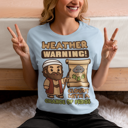 Light Blue t-shirt featuring a cute cartoon Moses pointing at a weather scroll that shows a smiling rain cloud and falling frogs. The text reads “Weather Warning! Cloudy With a Chance of Frogs.” The design uses playful retro colors with funny biblical humor inspired by the plague of frogs story