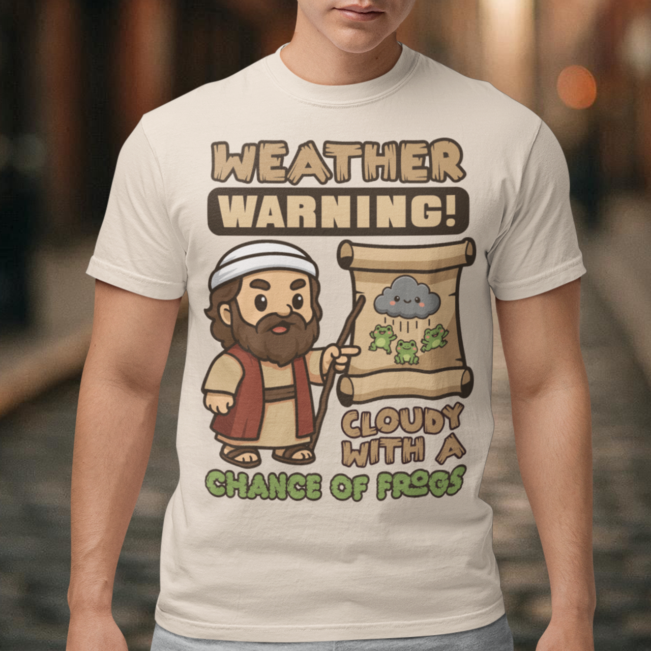 Beige t-shirt featuring a cute cartoon Moses pointing at a weather scroll that shows a smiling rain cloud and falling frogs. The text reads “Weather Warning! Cloudy With a Chance of Frogs.” The design uses playful retro colors with funny biblical humor inspired by the plague of frogs story