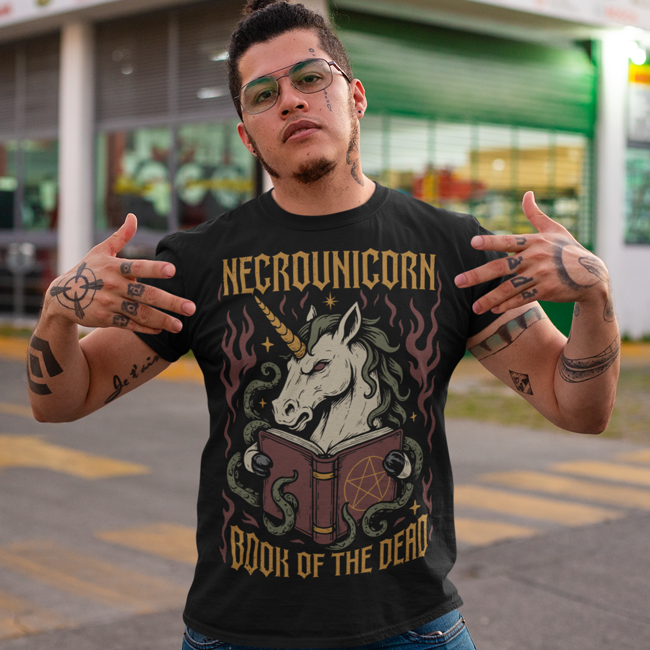 Black t-shirt featuring a dark fantasy graphic of an evil unicorn reading a spellbook with a pentagram symbol. The design includes flames, tentacles, stars, and gothic lettering reading “Necrounicorn” and “Book of the Dead.” Spooky occult-inspired artwork with horror, witchcore, and fantasy aesthetics.
