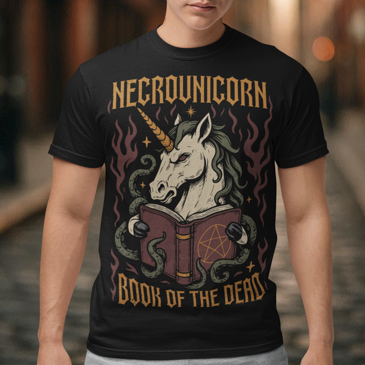 Black t-shirt featuring a dark fantasy graphic of an evil unicorn reading a spellbook with a pentagram symbol. The design includes flames, tentacles, stars, and gothic lettering reading “Necrounicorn” and “Book of the Dead.” Spooky occult-inspired artwork with horror, witchcore, and fantasy aesthetics.