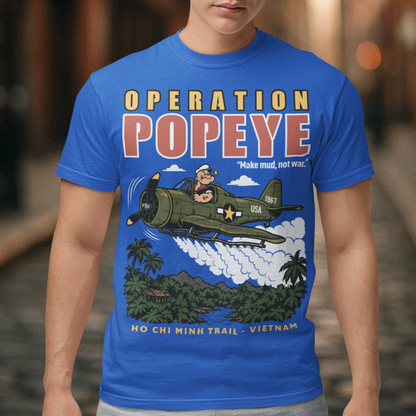 Blue T-shirt featuring a retro cartoon-style Popeye flying a vintage military aircraft over a lush Vietnam jungle landscape. The design reads “OPERATION POPEYE” with the phrase “Make mud, not war” and “HO CHI MINH TRAIL - VIETNAM.” A bold graphic inspired by Vietnam War history, aviation, military culture, and classic cartoon art.