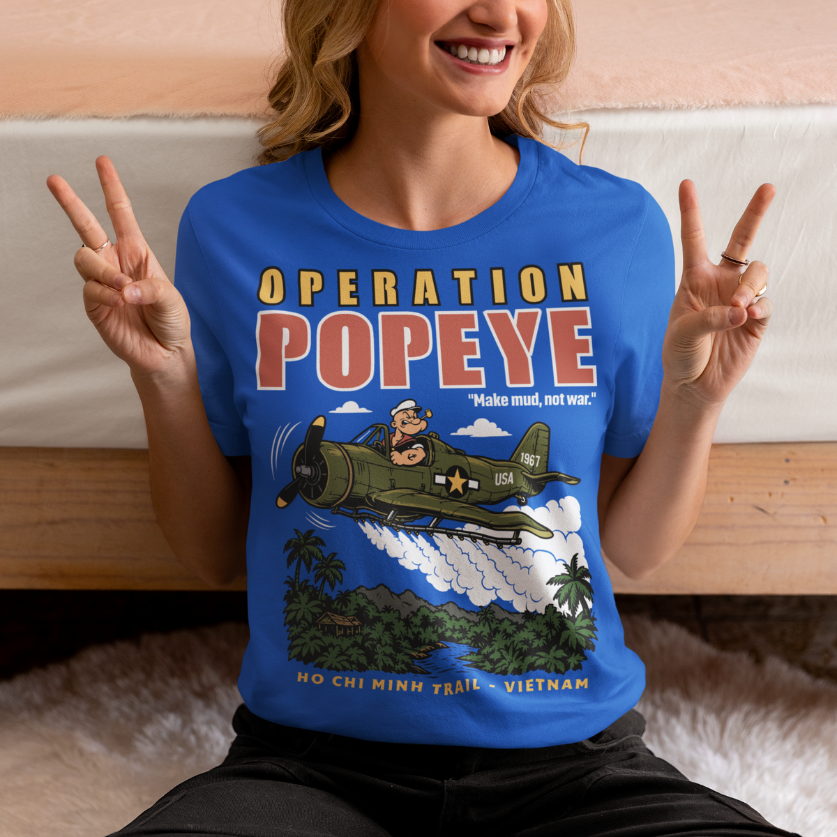 Blue T-shirt featuring a retro cartoon-style Popeye flying a vintage military aircraft over a lush Vietnam jungle landscape. The design reads “OPERATION POPEYE” with the phrase “Make mud, not war” and “HO CHI MINH TRAIL - VIETNAM.” A bold graphic inspired by Vietnam War history, aviation, military culture, and classic cartoon art.