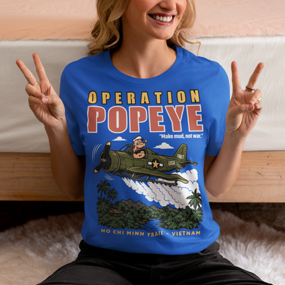 Blue T-shirt featuring a retro cartoon-style Popeye flying a vintage military aircraft over a lush Vietnam jungle landscape. The design reads “OPERATION POPEYE” with the phrase “Make mud, not war” and “HO CHI MINH TRAIL - VIETNAM.” A bold graphic inspired by Vietnam War history, aviation, military culture, and classic cartoon art.