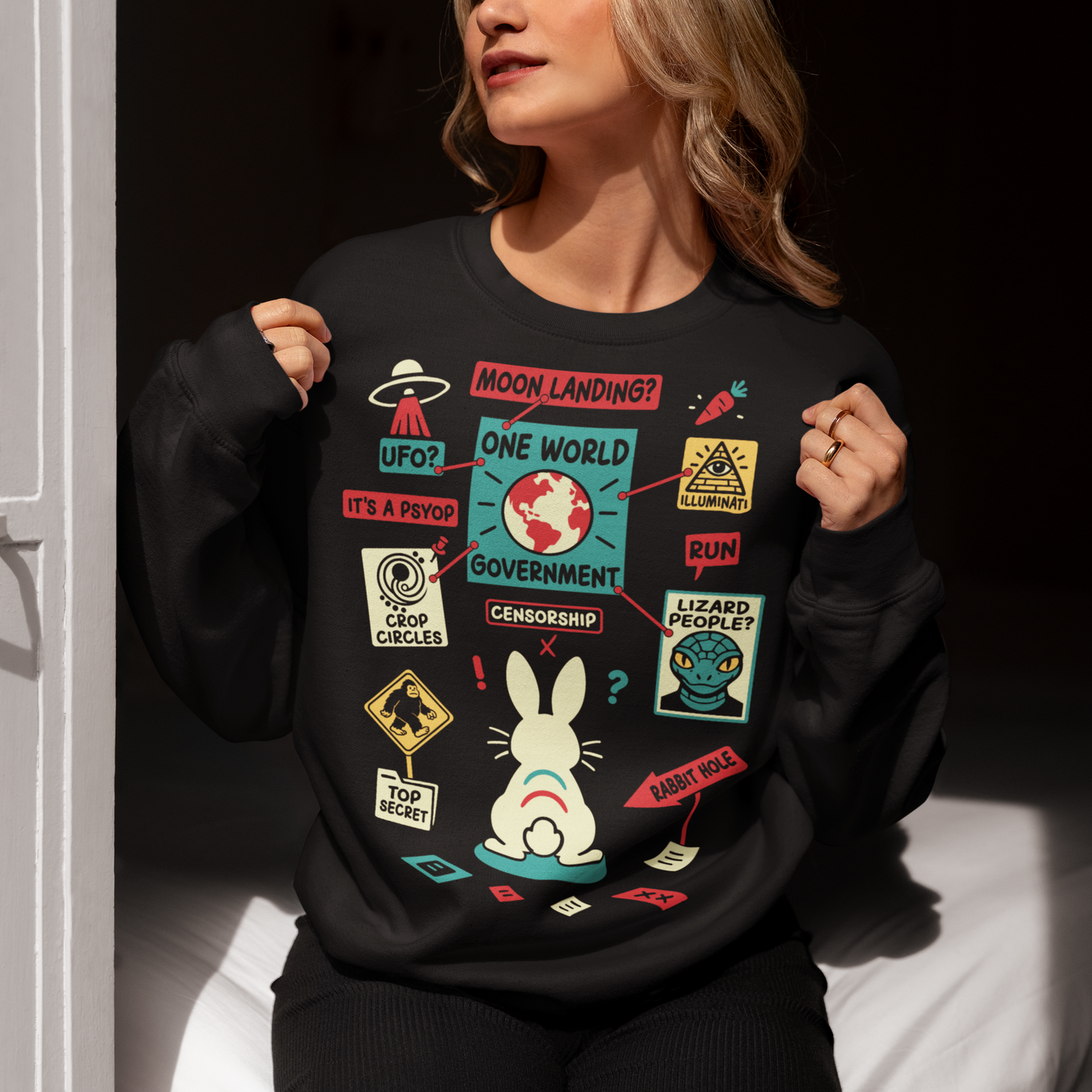 Black sweatshirt featuring a humorous conspiracy theory design with a confused rabbit standing before a bulletin board connected by red strings. The artwork includes UFOs, moon landing, lizard people, crop circles, Bigfoot, Illuminati, censorship, psyops, and rabbit hole references in a colorful retro style. A funny graphic tee for fans of conspiracies, cryptids, aliens, internet memes, and mystery-themed humor.