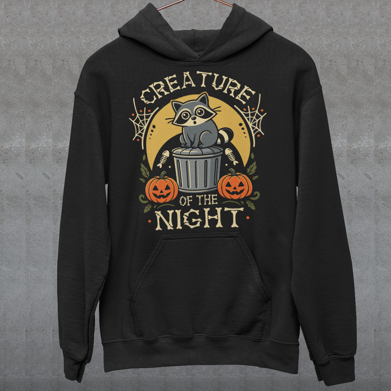 Black hoodie featuring a vintage Halloween graphic of a cute raccoon sitting on a trash can beneath a golden moon. The design includes pumpkins, spider webs, leaves, fish bones, and distressed lettering reading “CREATURE OF THE NIGHT.” A spooky woodland animal design perfect for raccoon lovers, Halloween fans, and anyone who loves nocturnal creatures.