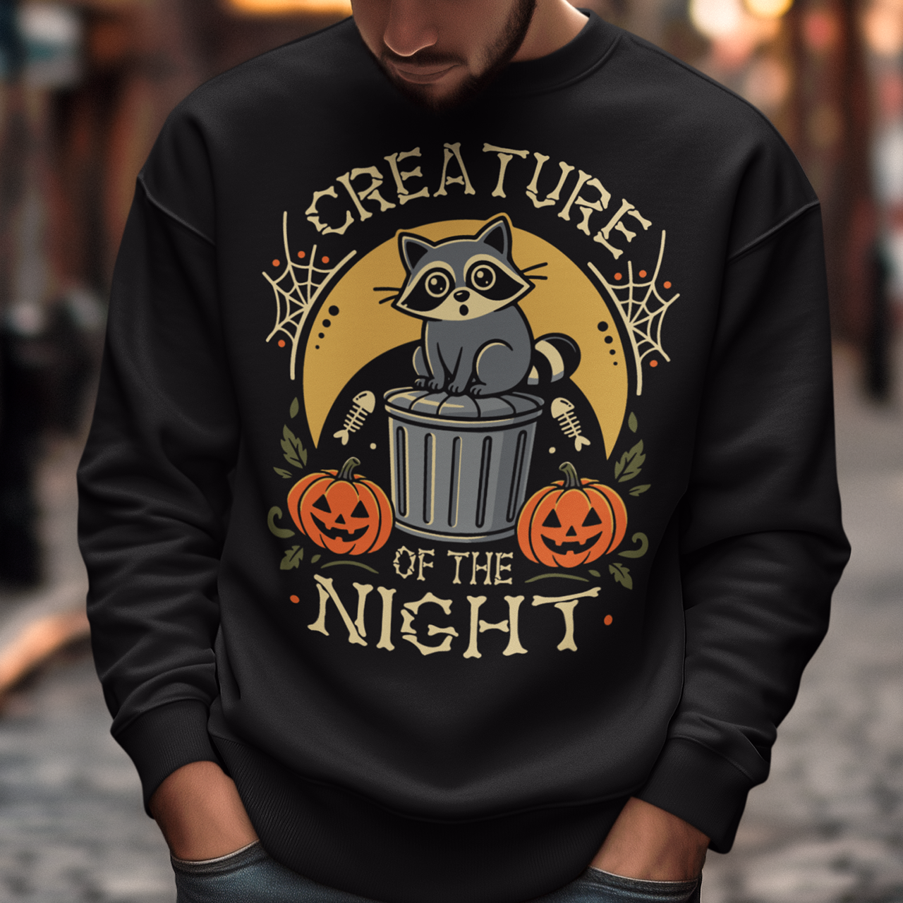 Black sweatshirt featuring a vintage Halloween graphic of a cute raccoon sitting on a trash can beneath a golden moon. The design includes pumpkins, spider webs, leaves, fish bones, and distressed lettering reading “CREATURE OF THE NIGHT.” A spooky woodland animal design perfect for raccoon lovers, Halloween fans, and anyone who loves nocturnal creatures.