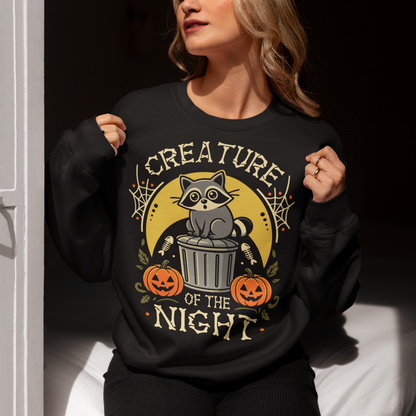 Black sweatshirt featuring a vintage Halloween graphic of a cute raccoon sitting on a trash can beneath a golden moon. The design includes pumpkins, spider webs, leaves, fish bones, and distressed lettering reading “CREATURE OF THE NIGHT.” A spooky woodland animal design perfect for raccoon lovers, Halloween fans, and anyone who loves nocturnal creatures.