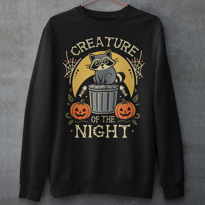 Black sweatshirt featuring a vintage Halloween graphic of a cute raccoon sitting on a trash can beneath a golden moon. The design includes pumpkins, spider webs, leaves, fish bones, and distressed lettering reading “CREATURE OF THE NIGHT.” A spooky woodland animal design perfect for raccoon lovers, Halloween fans, and anyone who loves nocturnal creatures.