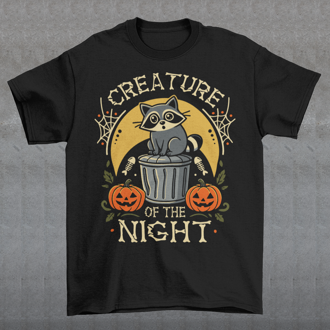 Black t-shirt featuring a vintage Halloween graphic of a cute raccoon sitting on a trash can beneath a golden moon. The design includes pumpkins, spider webs, leaves, fish bones, and distressed lettering reading “CREATURE OF THE NIGHT.” A spooky woodland animal design perfect for raccoon lovers, Halloween fans, and anyone who loves nocturnal creatures.
