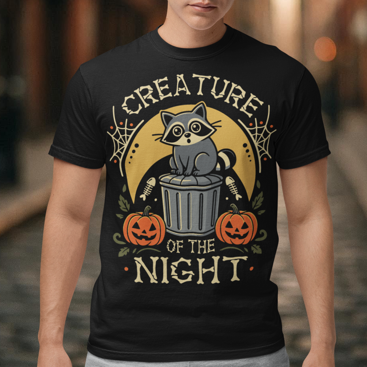 Black t-shirt featuring a vintage Halloween graphic of a cute raccoon sitting on a trash can beneath a golden moon. The design includes pumpkins, spider webs, leaves, fish bones, and distressed lettering reading “CREATURE OF THE NIGHT.” A spooky woodland animal design perfect for raccoon lovers, Halloween fans, and anyone who loves nocturnal creatures.