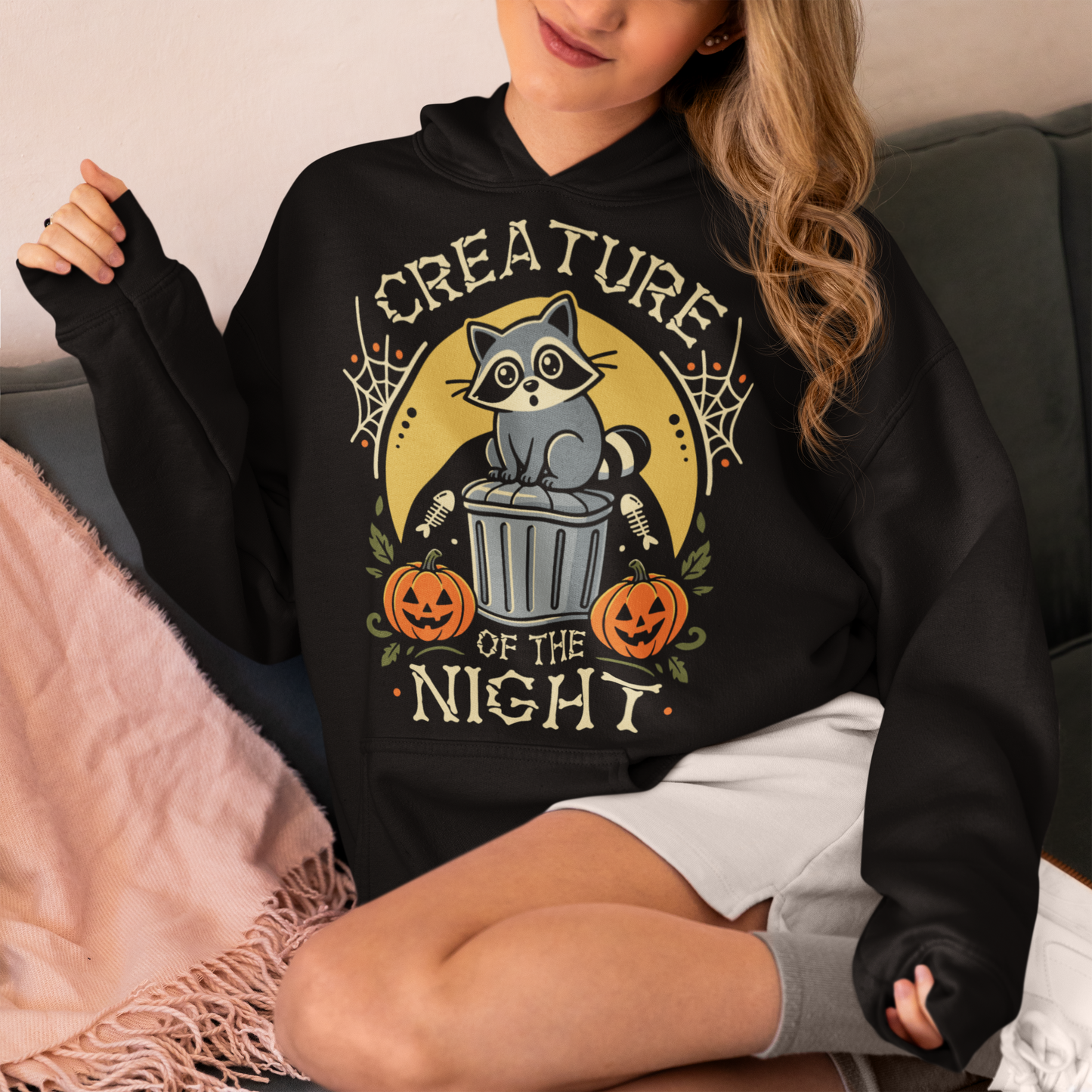 Black hoodie featuring a vintage Halloween graphic of a cute raccoon sitting on a trash can beneath a golden moon. The design includes pumpkins, spider webs, leaves, fish bones, and distressed lettering reading “CREATURE OF THE NIGHT.” A spooky woodland animal design perfect for raccoon lovers, Halloween fans, and anyone who loves nocturnal creatures.