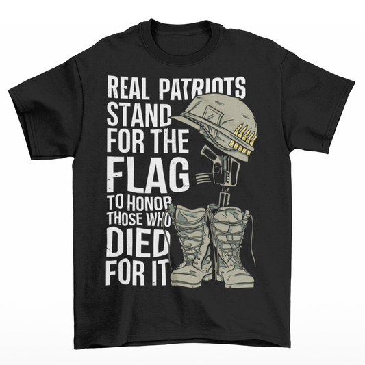 -shirt design featuring a graphic of a military helmet resting on a rifle, which is standing upright in a pair of combat boots—a battlefield cross symbolizing fallen soldiers. Above and below the image is bold text that reads: "REAL PATRIOTS STAND FOR THE FLAG TO HONOR THOSE WHO DIED FOR IT." The design uses patriotic imagery to emphasize respect and remembrance for military service members who have sacrificed their lives.