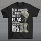 -shirt design featuring a graphic of a military helmet resting on a rifle, which is standing upright in a pair of combat boots—a battlefield cross symbolizing fallen soldiers. Above and below the image is bold text that reads: "REAL PATRIOTS STAND FOR THE FLAG TO HONOR THOSE WHO DIED FOR IT." The design uses patriotic imagery to emphasize respect and remembrance for military service members who have sacrificed their lives.
