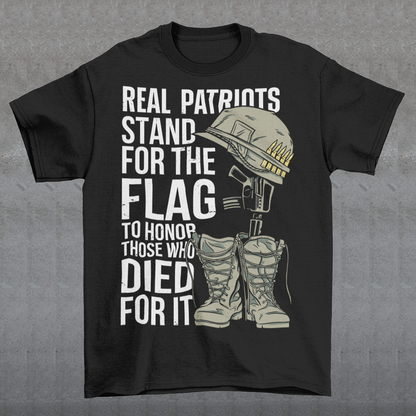 -shirt design featuring a graphic of a military helmet resting on a rifle, which is standing upright in a pair of combat boots—a battlefield cross symbolizing fallen soldiers. Above and below the image is bold text that reads: "REAL PATRIOTS STAND FOR THE FLAG TO HONOR THOSE WHO DIED FOR IT." The design uses patriotic imagery to emphasize respect and remembrance for military service members who have sacrificed their lives.