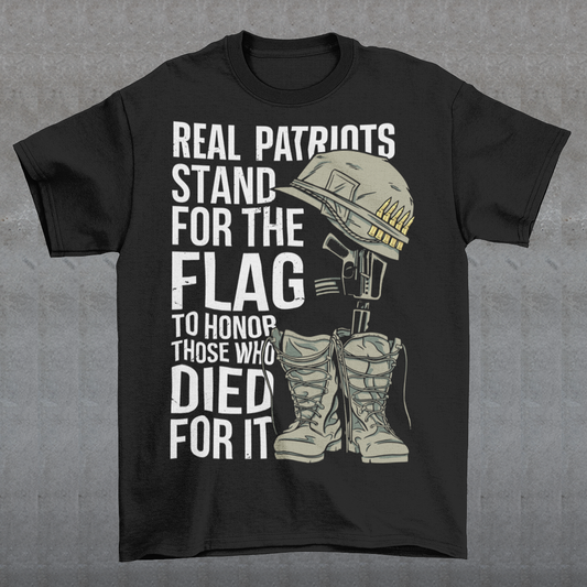 -shirt design featuring a graphic of a military helmet resting on a rifle, which is standing upright in a pair of combat boots—a battlefield cross symbolizing fallen soldiers. Above and below the image is bold text that reads: "REAL PATRIOTS STAND FOR THE FLAG TO HONOR THOSE WHO DIED FOR IT." The design uses patriotic imagery to emphasize respect and remembrance for military service members who have sacrificed their lives.