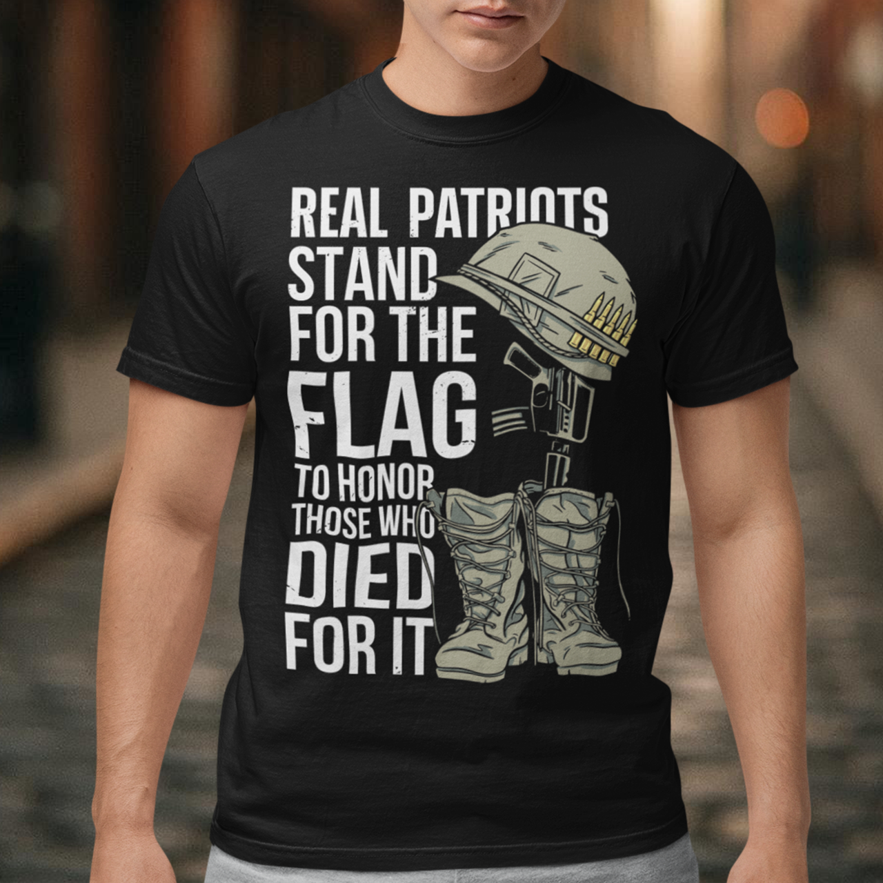 -shirt design featuring a graphic of a military helmet resting on a rifle, which is standing upright in a pair of combat boots—a battlefield cross symbolizing fallen soldiers. Above and below the image is bold text that reads: "REAL PATRIOTS STAND FOR THE FLAG TO HONOR THOSE WHO DIED FOR IT." The design uses patriotic imagery to emphasize respect and remembrance for military service members who have sacrificed their lives.