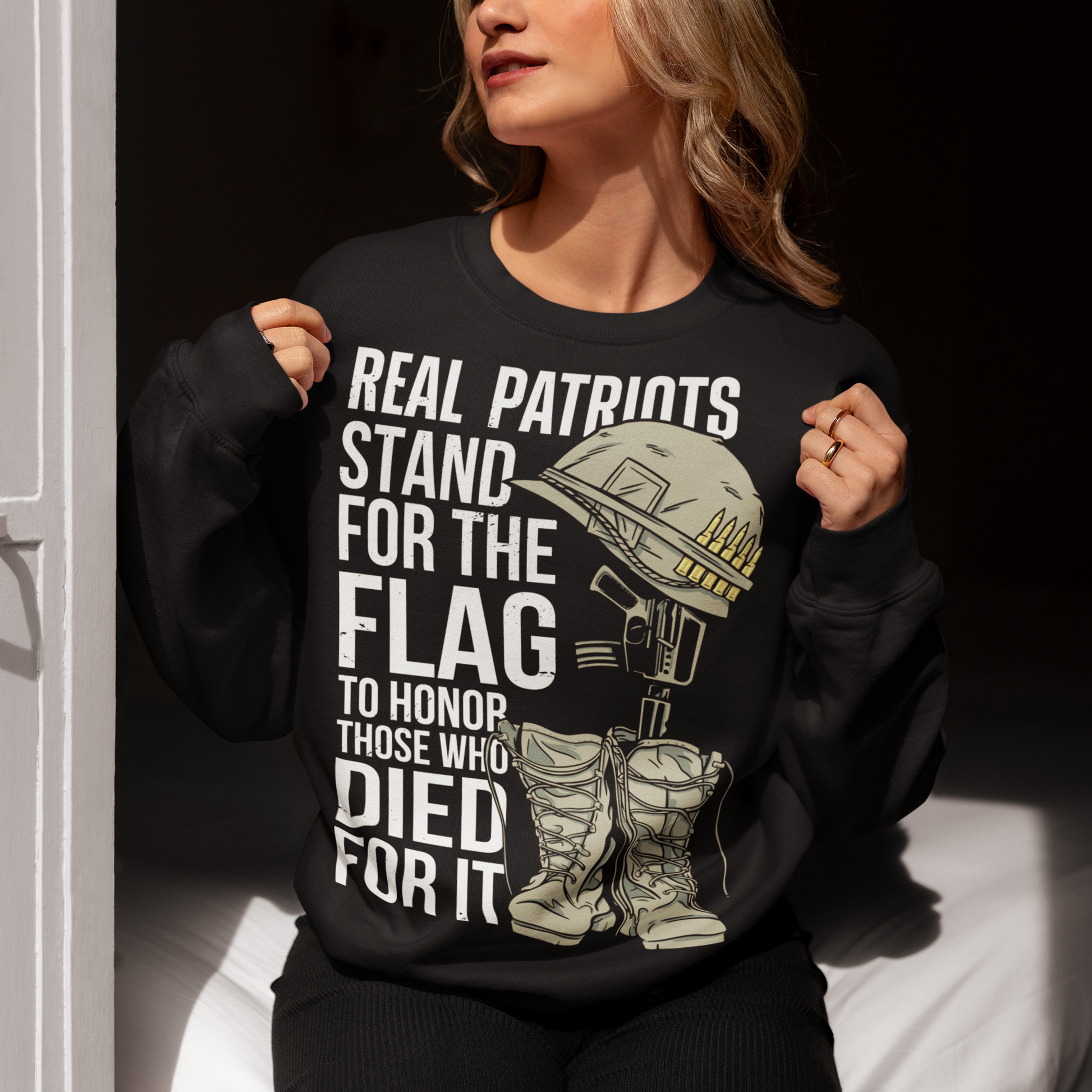 Sweatshirt design featuring a graphic of a military helmet resting on a rifle, which is standing upright in a pair of combat boots—a battlefield cross symbolizing fallen soldiers. Above and below the image is bold text that reads: "REAL PATRIOTS STAND FOR THE FLAG TO HONOR THOSE WHO DIED FOR IT." The design uses patriotic imagery to emphasize respect and remembrance for military service members who have sacrificed their lives.