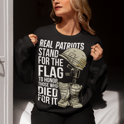 Sweatshirt design featuring a graphic of a military helmet resting on a rifle, which is standing upright in a pair of combat boots—a battlefield cross symbolizing fallen soldiers. Above and below the image is bold text that reads: "REAL PATRIOTS STAND FOR THE FLAG TO HONOR THOSE WHO DIED FOR IT." The design uses patriotic imagery to emphasize respect and remembrance for military service members who have sacrificed their lives.