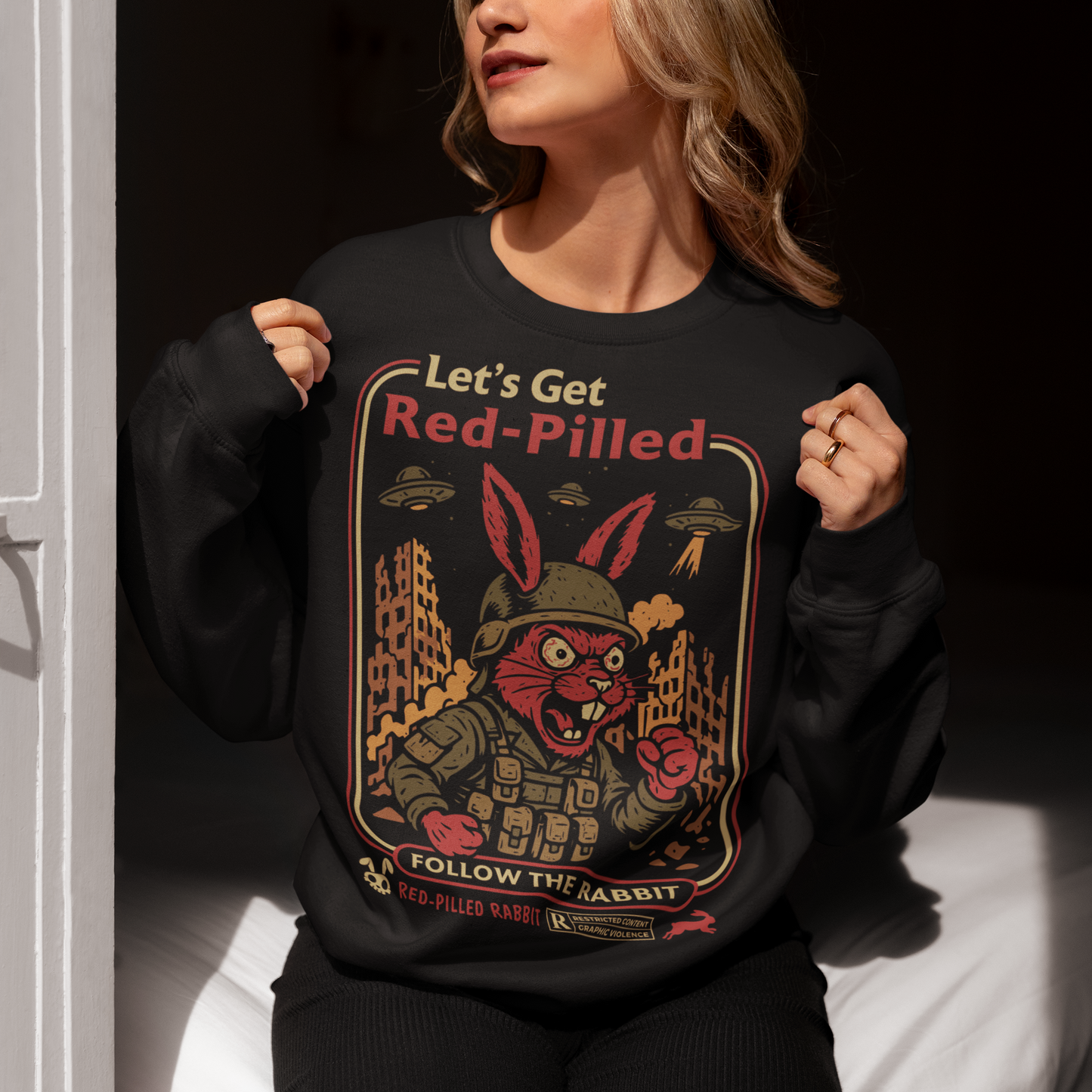 This black graphic sweatshirt dives headfirst down the rabbit hole. Featuring a fierce, red-eyed rabbit geared up like a rebel soldier, the design blends sci-fi paranoia, retro poster aesthetics, and counterculture attitude. Bold lettering reads “Let’s Get Red-Pilled” with “Follow the Rabbit” anchoring the scene, while crumbling cityscapes and hovering UFOs set a dystopian, mind-bending mood. This shirt feels like a lost propaganda poster from an alternate reality.