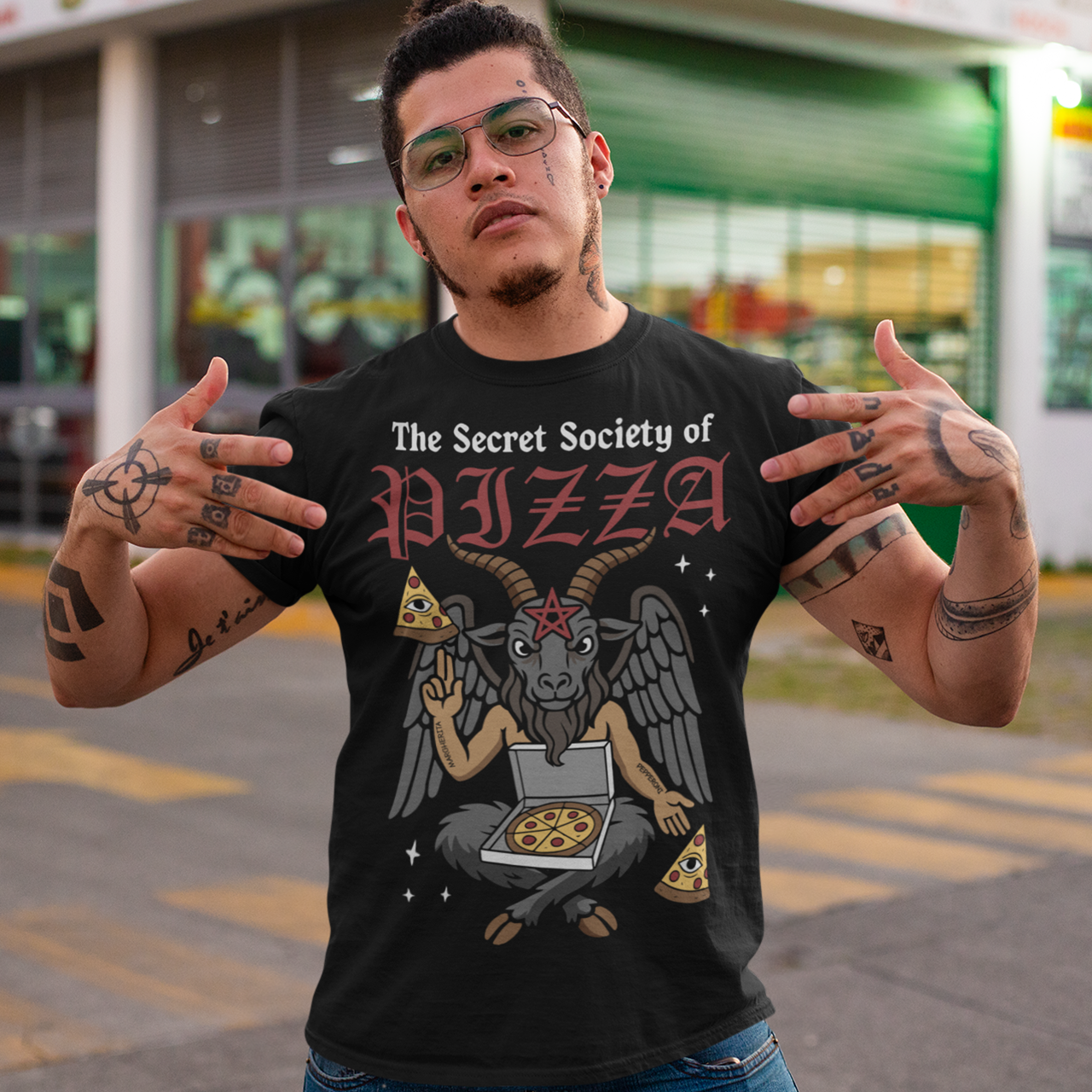 Black t-shirt featuring a humorous occult-style design with the text “The Secret Society of Pizza.” Artwork shows a winged goat figure with horns and a pentagram holding pizza slices and an open pizza box, surrounded by small stars. Gothic lettering and playful dark humor create a spooky pizza-themed graphic tee design.