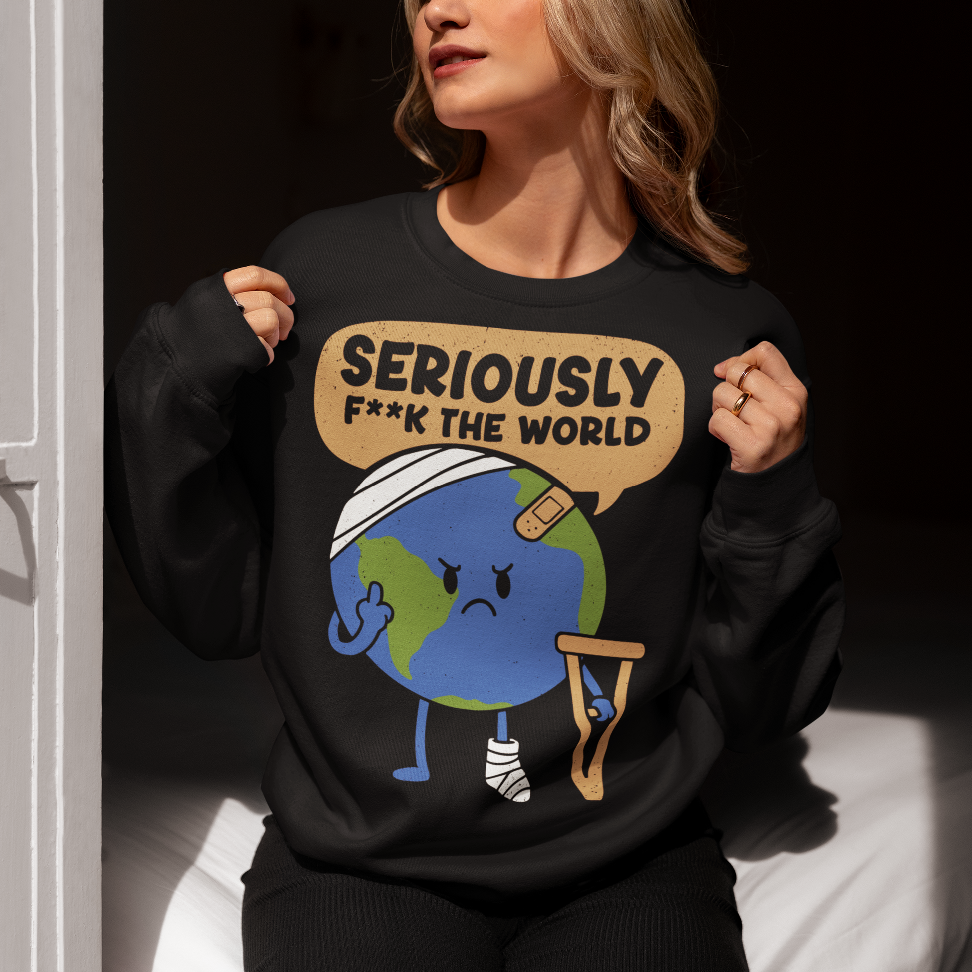 Black sweatshirt featuring a funny cartoon Earth character with bandages, crutches, and an angry face flipping the middle finger. Above the character is the text “Seriously F**k The World” in bold lettering. The design uses bright blue and green Earth colors against a black shirt for a dark humor, sarcastic graphic shirt style.