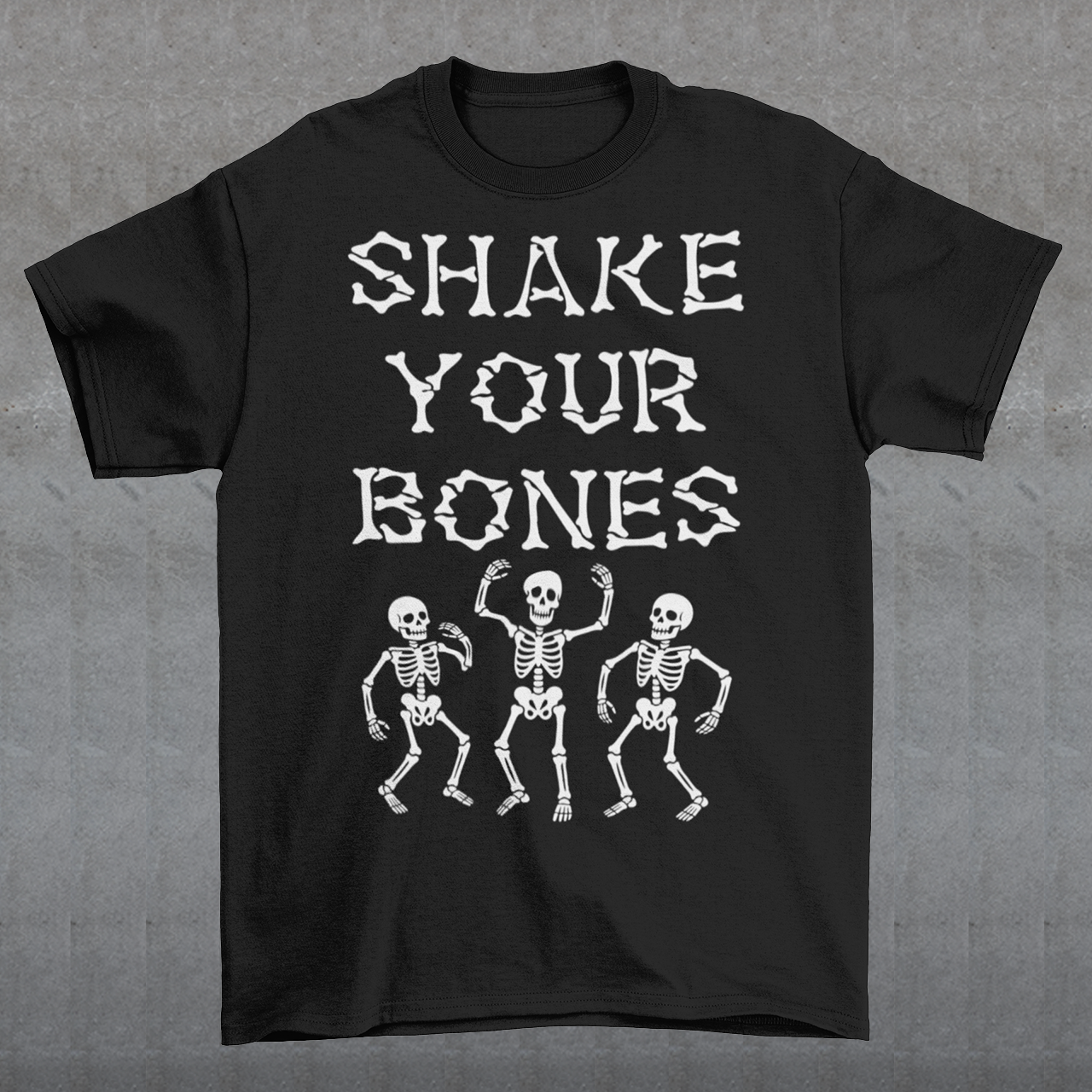 Black t-shirt featuring bold distressed lettering reading “SHAKE YOUR BONES” above three dancing skeletons. The vintage-inspired graphic uses white artwork on a black background, creating a spooky gothic look with dark humor. Perfect for Halloween, skeleton lovers, goth fashion fans, spooky season, parties, concerts, and anyone who likes their wardrobe with a little skeletal attitude.
