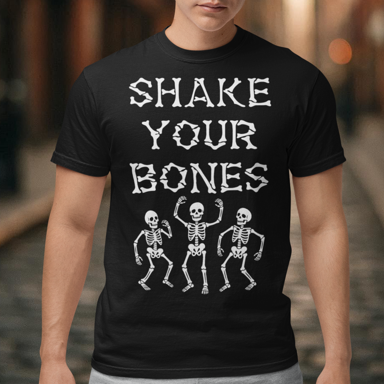 Black t-shirt featuring bold distressed lettering reading “SHAKE YOUR BONES” above three dancing skeletons. The vintage-inspired graphic uses white artwork on a black background, creating a spooky gothic look with dark humor. Perfect for Halloween, skeleton lovers, goth fashion fans, spooky season, parties, concerts, and anyone who likes their wardrobe with a little skeletal attitude.