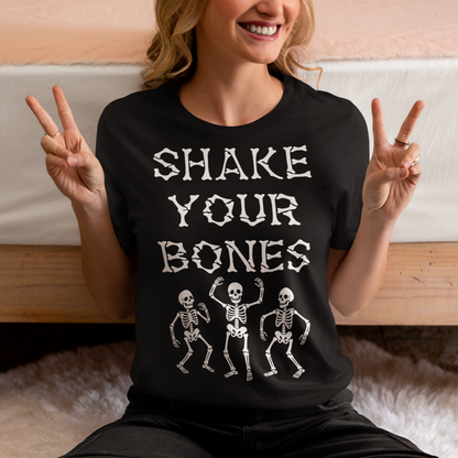 Black t-shirt featuring bold distressed lettering reading “SHAKE YOUR BONES” above three dancing skeletons. The vintage-inspired graphic uses white artwork on a black background, creating a spooky gothic look with dark humor. Perfect for Halloween, skeleton lovers, goth fashion fans, spooky season, parties, concerts, and anyone who likes their wardrobe with a little skeletal attitude.