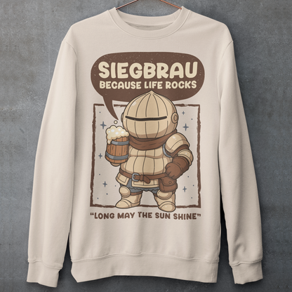Beige sweatshirt featuring a cute, chibi-style armored knight Siegmeyer of Catarina holding a frothy beer mug. Above the character, bold text reads “Siegbrau because life rocks,” with smaller text below saying “Long may the sun shine.” The design has a playful fantasy theme with soft brown tones and a hand-drawn style, appealing to gamers and fans of medieval or RPG-inspired humor.