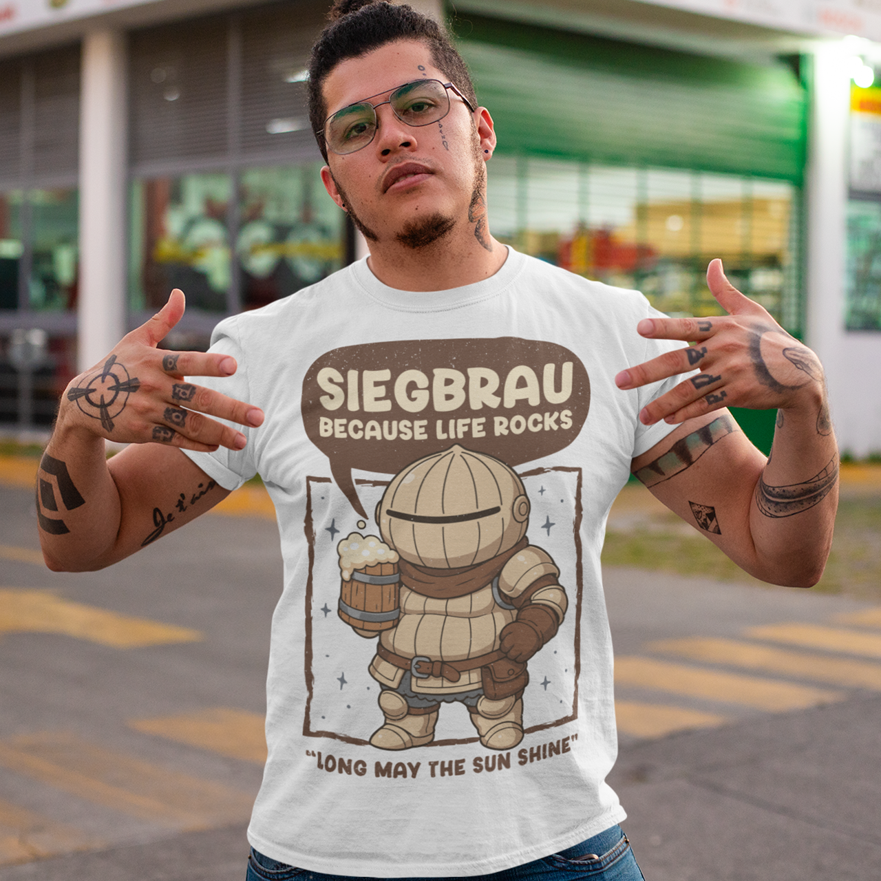 White t-shirt featuring a cute, chibi-style armored knight Siegmeyer of Catarina holding a frothy beer mug. Above the character, bold text reads “Siegbrau because life rocks,” with smaller text below saying “Long may the sun shine.” The design has a playful fantasy theme with soft brown tones and a hand-drawn style, appealing to gamers and fans of medieval or RPG-inspired humor.