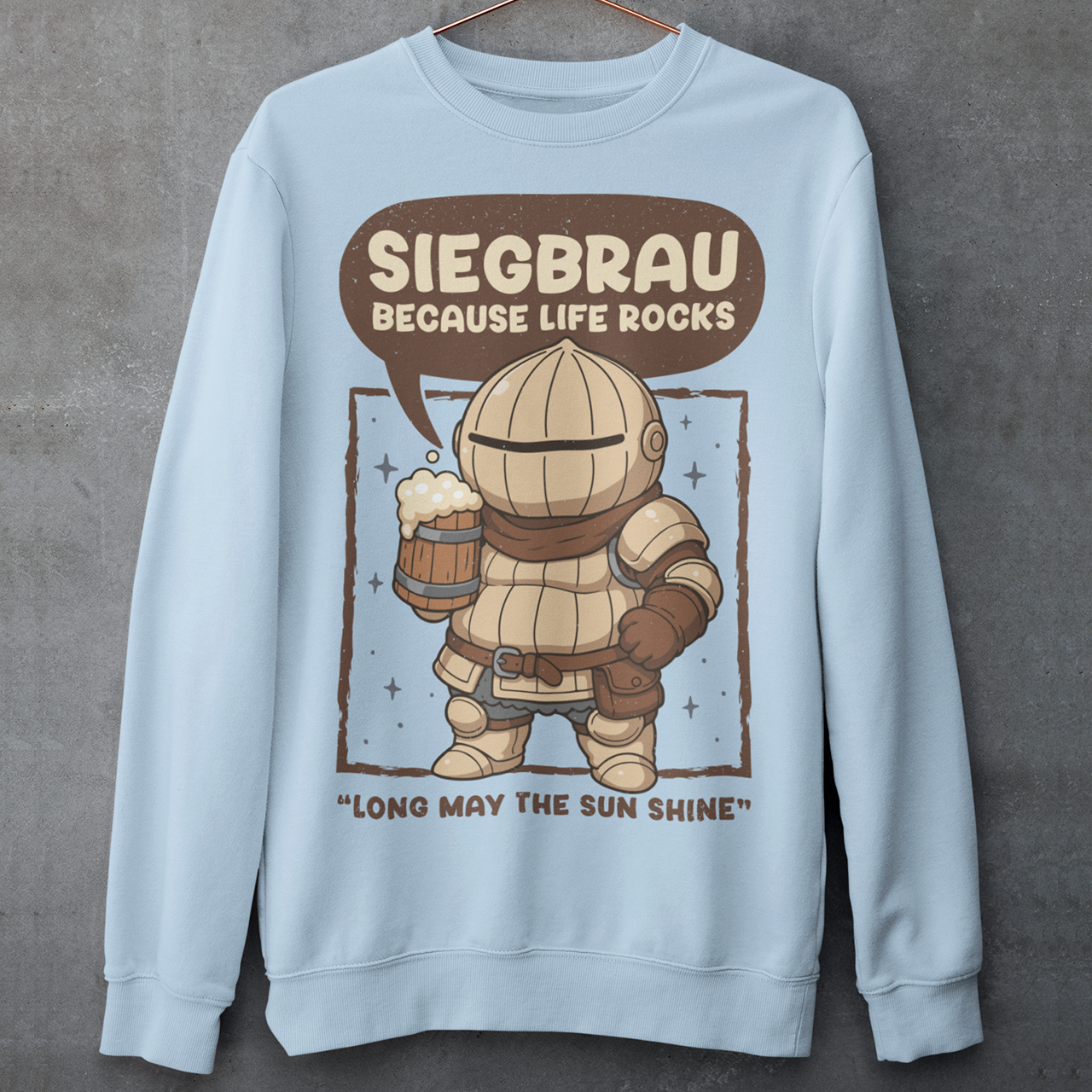 Light Blue sweatshirt featuring a cute, chibi-style armored knight Siegmeyer of Catarina holding a frothy beer mug. Above the character, bold text reads “Siegbrau because life rocks,” with smaller text below saying “Long may the sun shine.” The design has a playful fantasy theme with soft brown tones and a hand-drawn style, appealing to gamers and fans of medieval or RPG-inspired humor.