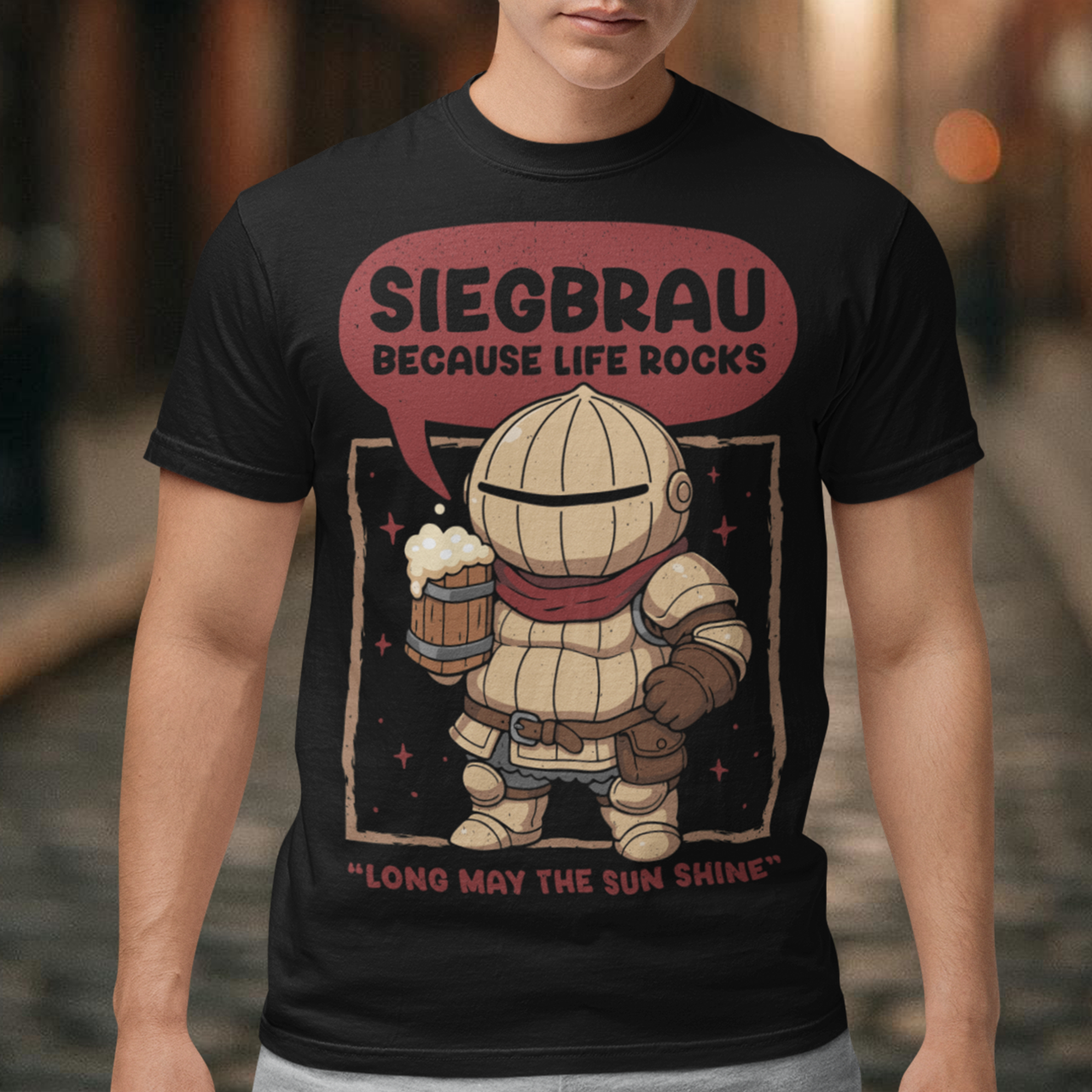 Black t-shirt featuring a cute, chibi-style armored knight Siegmeyer of Catarina holding a frothy beer mug. Above the character, bold text reads “Siegbrau because life rocks,” with smaller text below saying “Long may the sun shine.” The design has a playful fantasy theme with soft red tones and a hand-drawn style, appealing to gamers and fans of medieval or RPG-inspired humor.