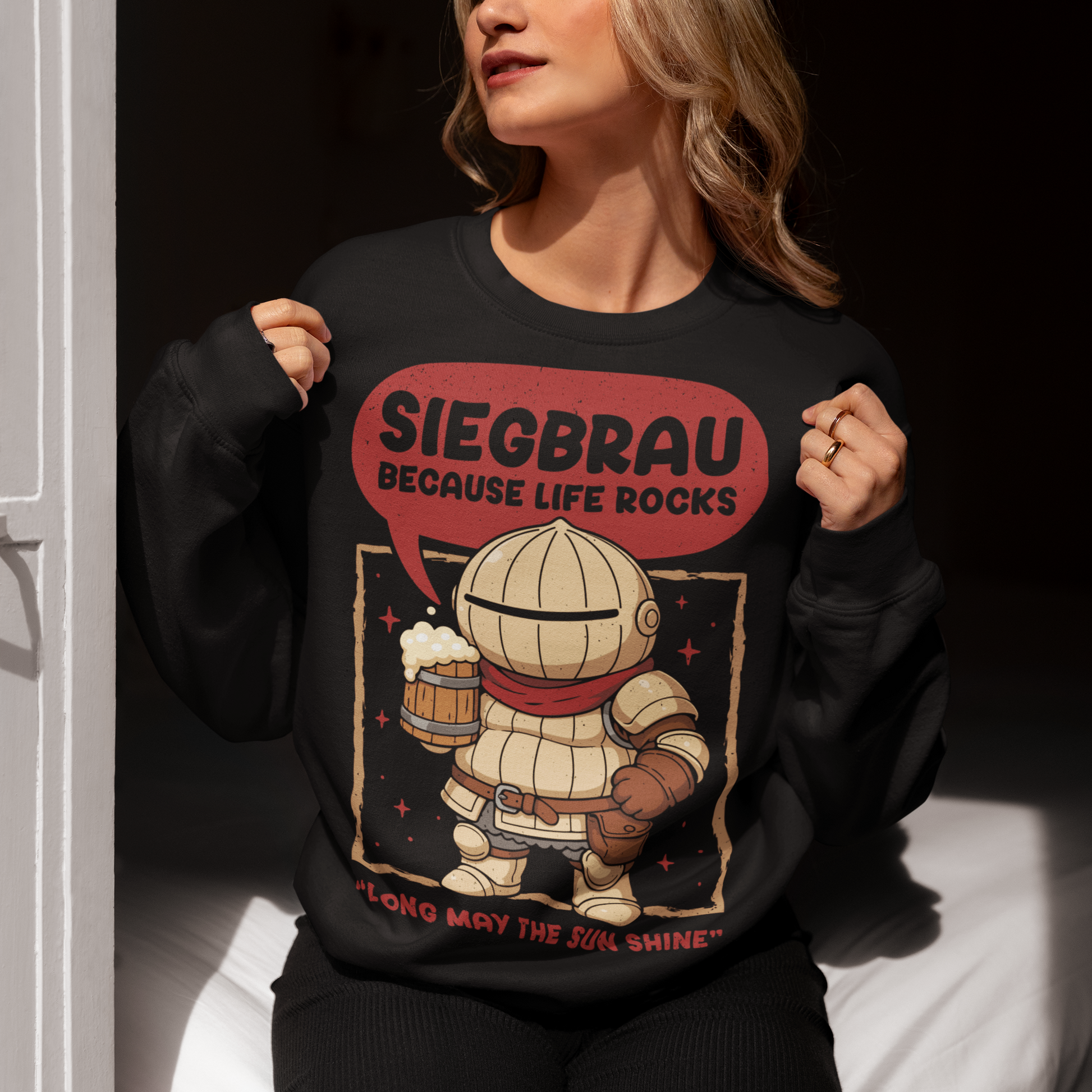 Black sweatshirt featuring a cute, chibi-style armored knight Siegmeyer of Catarina holding a frothy beer mug. Above the character, bold text reads “Siegbrau because life rocks,” with smaller text below saying “Long may the sun shine.” The design has a playful fantasy theme with soft red tones and a hand-drawn style, appealing to gamers and fans of medieval or RPG-inspired humor.