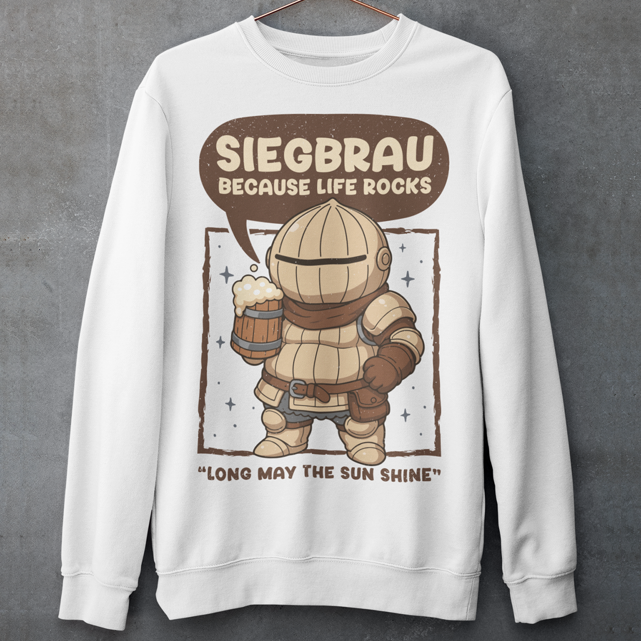 White sweatshirt featuring a cute, chibi-style armored knight Siegmeyer of Catarina holding a frothy beer mug. Above the character, bold text reads “Siegbrau because life rocks,” with smaller text below saying “Long may the sun shine.” The design has a playful fantasy theme with soft brown tones and a hand-drawn style, appealing to gamers and fans of medieval or RPG-inspired humor.